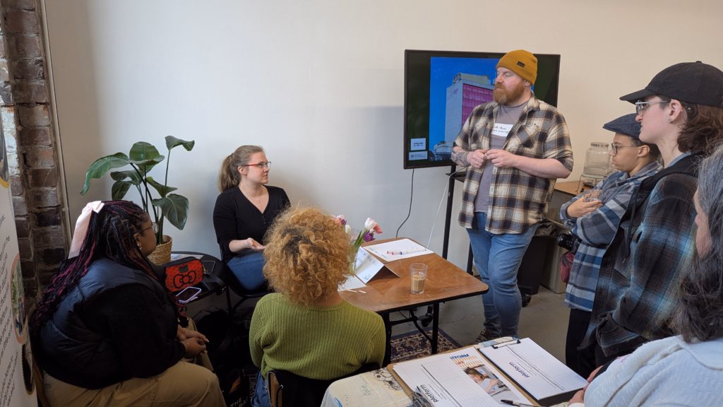 A small group discusses around a table with a laptop screen displaying an image, surrounded by plants and cheerful decor.
