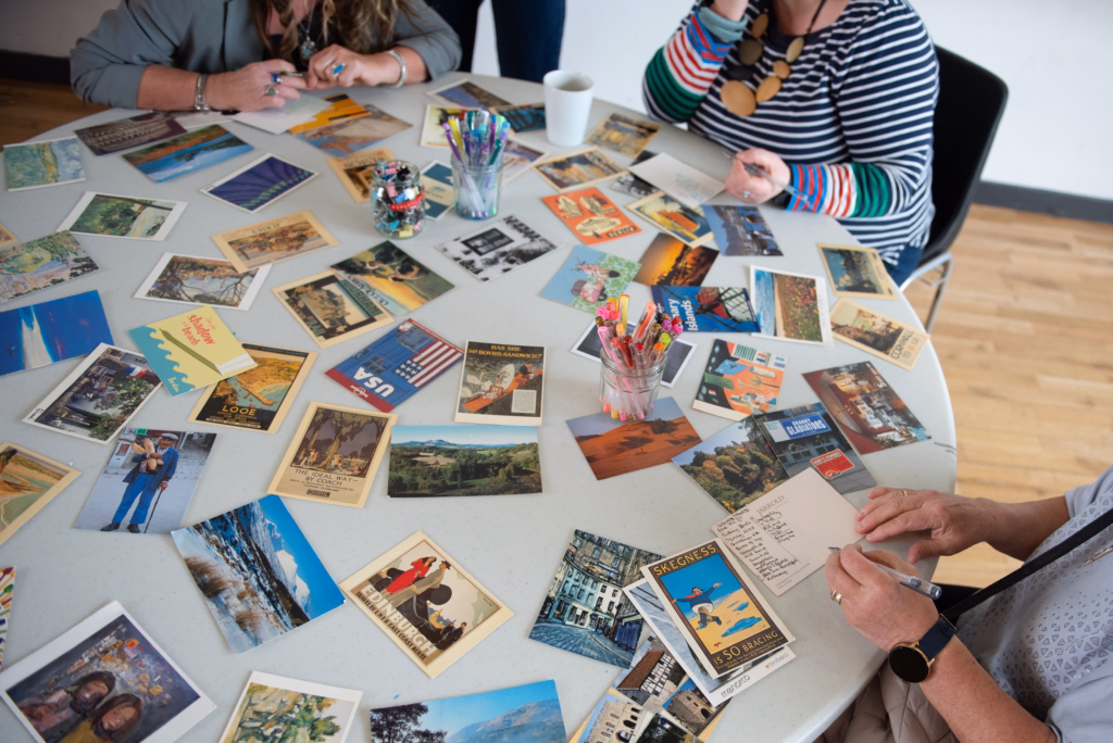 Three people sit at a round table covered with postcards, pens, and colored pencils, each writing on a postcard.