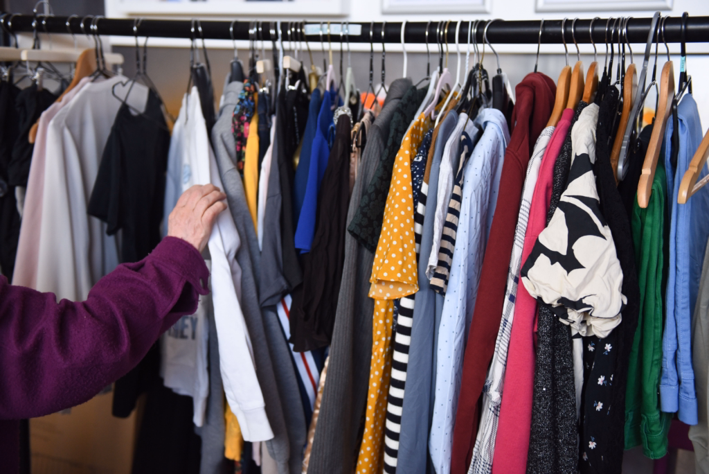 A person in a purple sweater reaches for a colourful array of hanging shirts on a clothing rack in a well-lit room.
