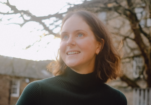 Jo is looking away from the camera smiling. She is outside with a tree behind her. She has shoulder length dark hair and a green polo neck top on